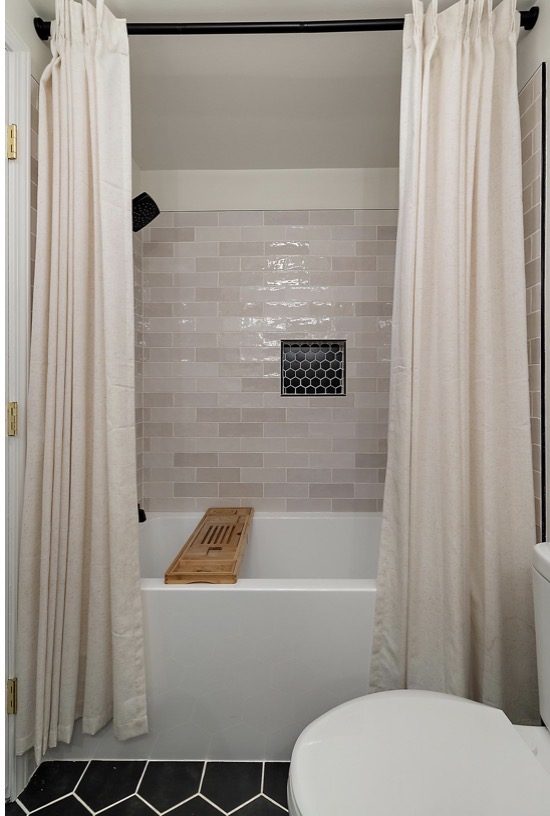 Tub and shower remodel with light gray subway tile, black hexagon niche, and linen curtains — Downriver Michigan